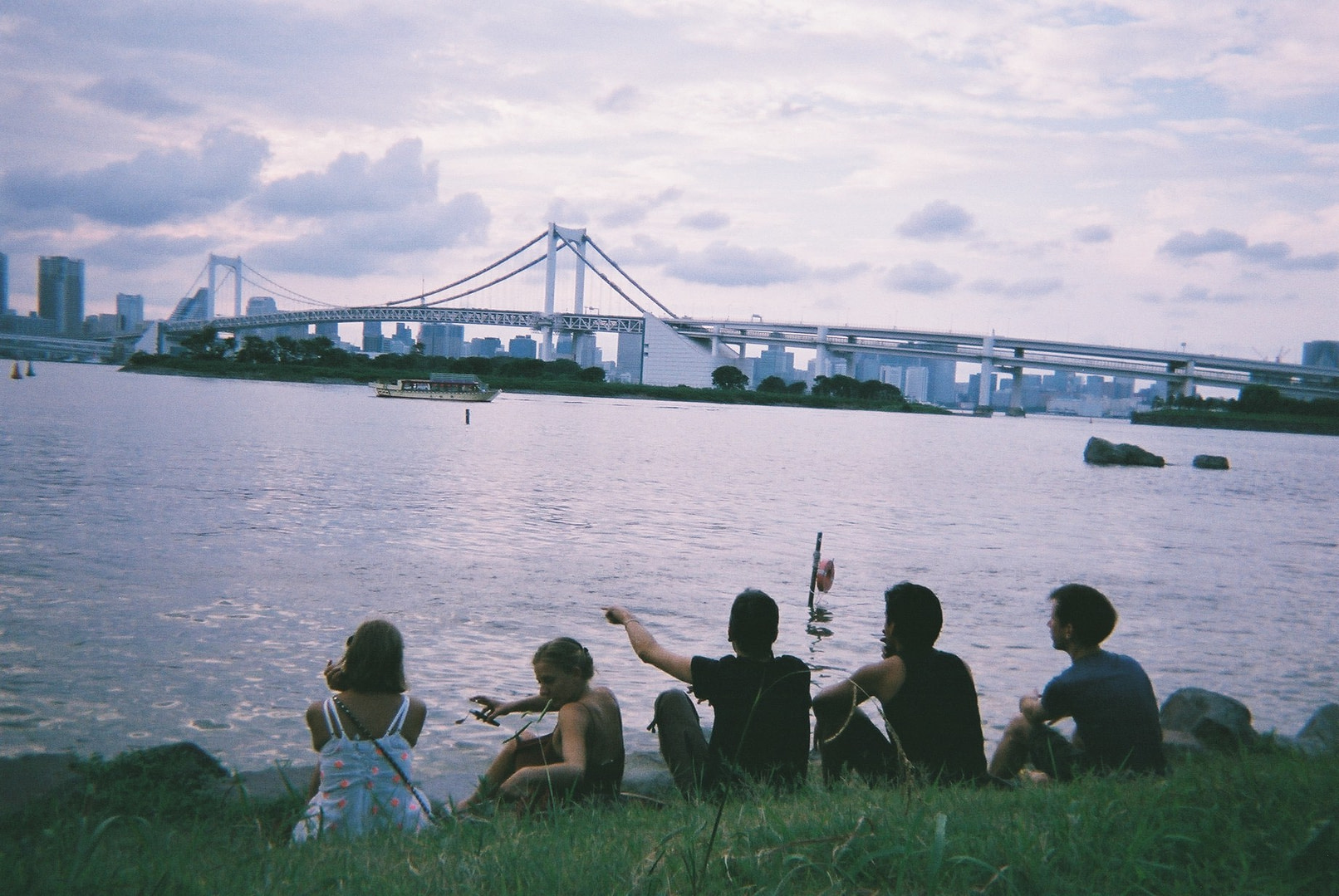 five young adults sitting in a patch of grass in front of a body of water and a view of the city in the distance on a cloudy day