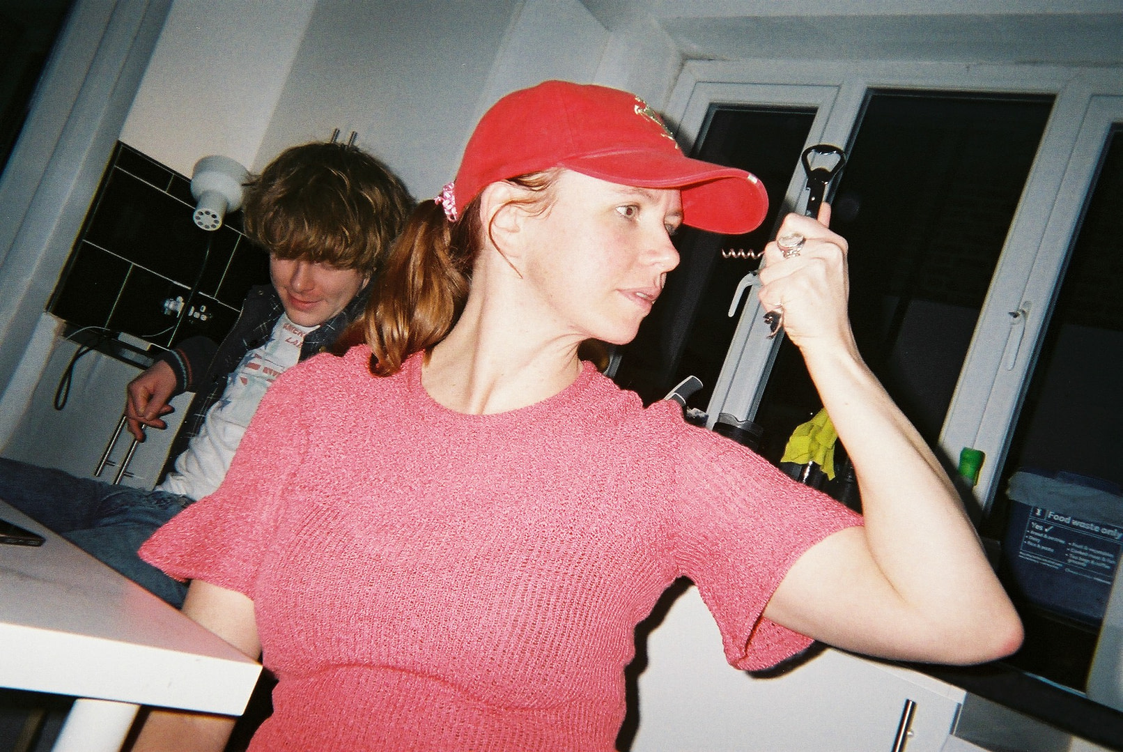 a woman wearing a red cap and a pink top with a corkscew in her hand directed towards herself, in a kitchen