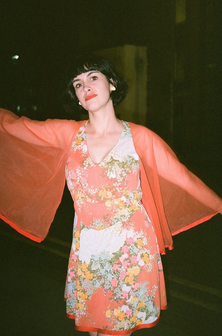 B from superorganism wearing an orange floral dress with sheer flowy sleeves, arms spread out