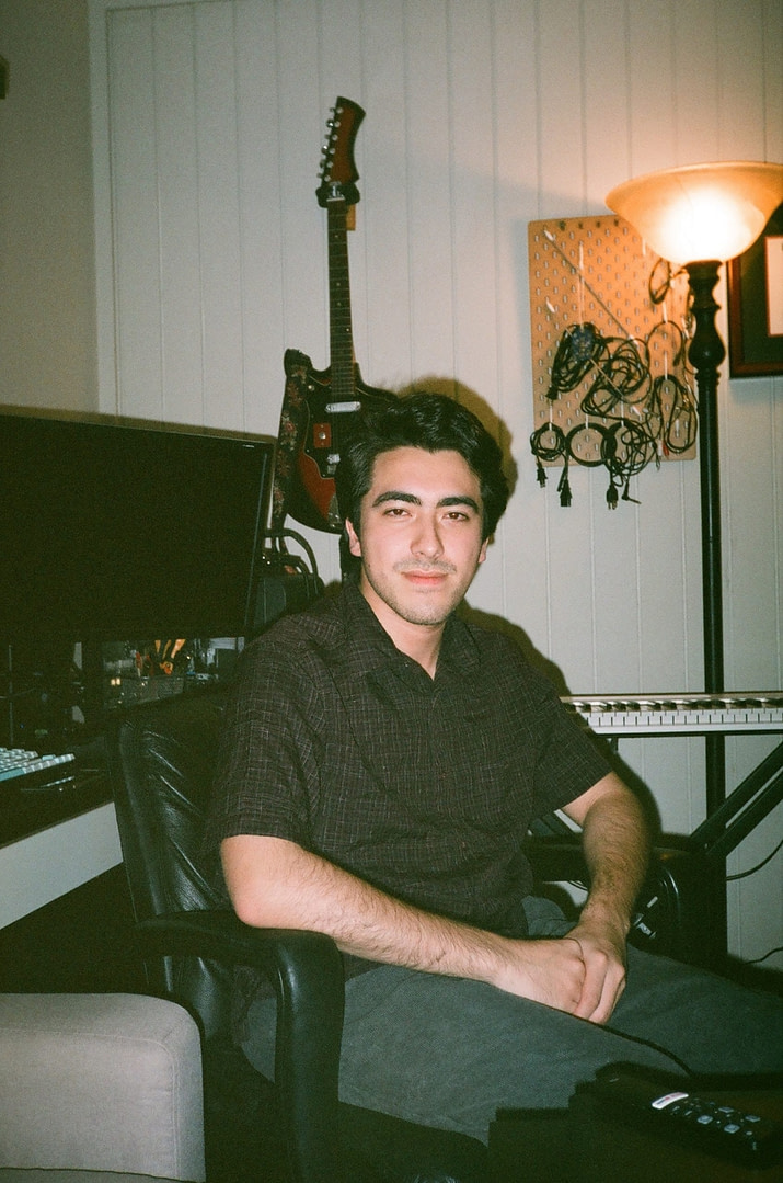 a boy sitting in his studio looking towards the camera, guitar, keyboard, and other recording equipment in the background